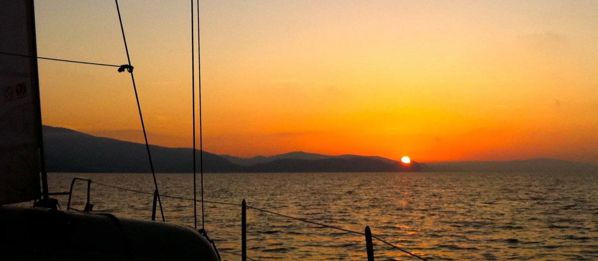 Alba in mare vista dal ponte di una barca a vela: la randa e le sartie in controluce sulla sinistra, la prua e le draglie in basso, il sole appena sopra il profilo dell'Isola d'Elba all'orizzonte e il cielo che passa dall'arancio al grigio azzurro
